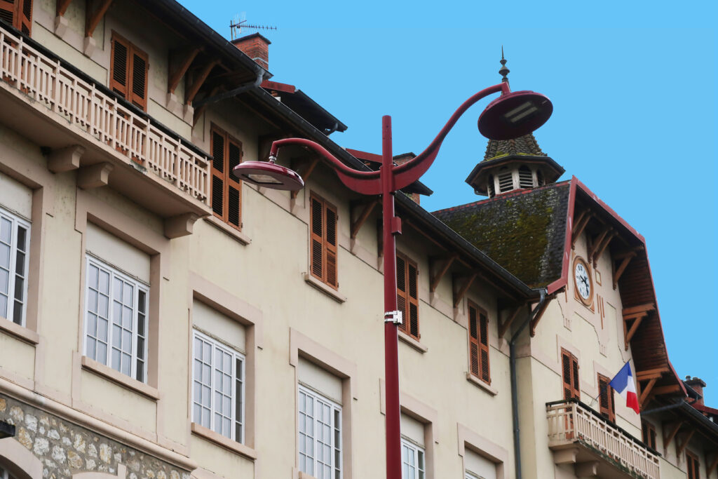 Bâtiment à la facade blanche, avec un balcon des fenêtre à double battant blanche au premier étage et des fenêtres rouges au second. Au centre du bâtiment on voit un drapeau francais et une horloge à cadran. Au premier plan un lampadaire rouge est visible.  - Agrandir l'image, fenêtre modale