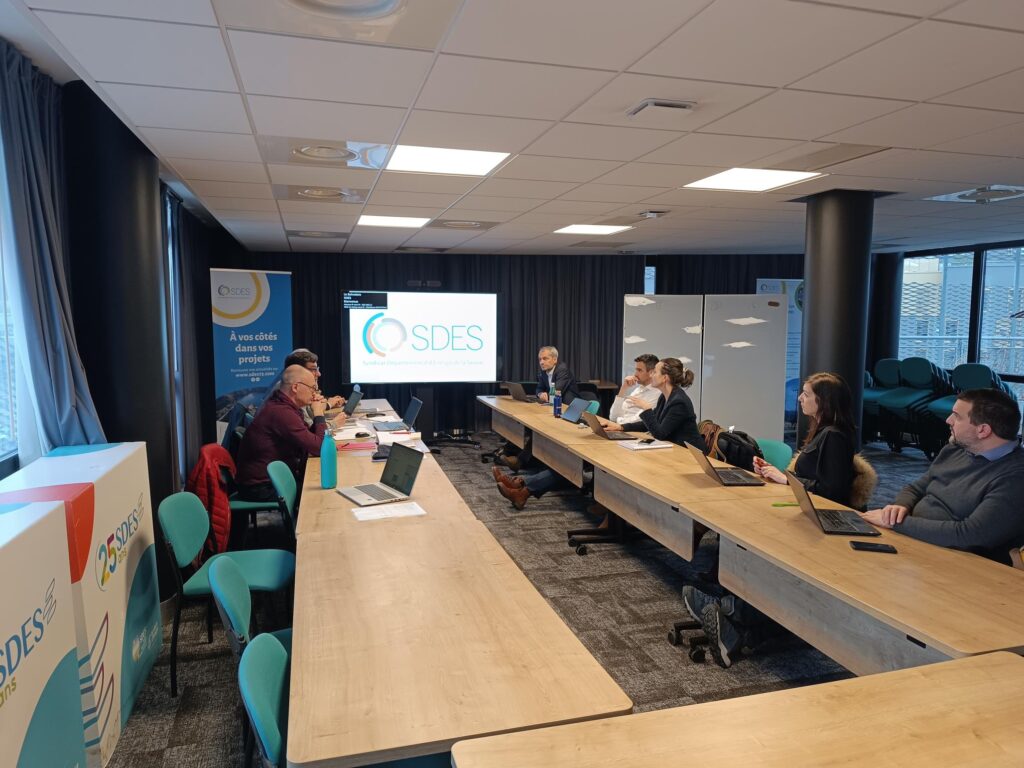 Salle de réunion, plutôt sombre avec une moquette grise et des rideaux noir recouvrants les baies vitrées. 
Autour de tables de réunis en U, huit personnes, devant des ordinateurs, parlent entre eux.  - Agrandir l'image, fenêtre modale