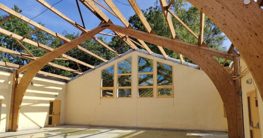 Photo de la salle polyvalente en rénovation ave des murs bruts, quatre grandes fenêtres et une charpente de toit en bois visibles et nu. 
À travers la charpente, on aperçoit un ciel bleu, sans nuage et le sommet de plusieurs arbres verts.  - Agrandir l'image, fenêtre modale